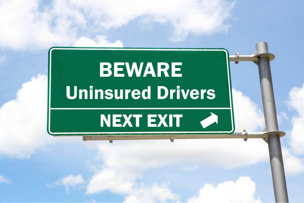 Green overhead road sign with a Beware of Uninsured Drivers Next Exit concept against a partly cloudy sky background.